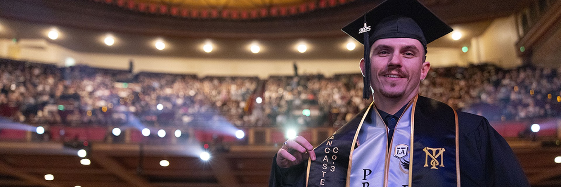 A student wearing graduation attire smiles, an auditorium full of people behind him.