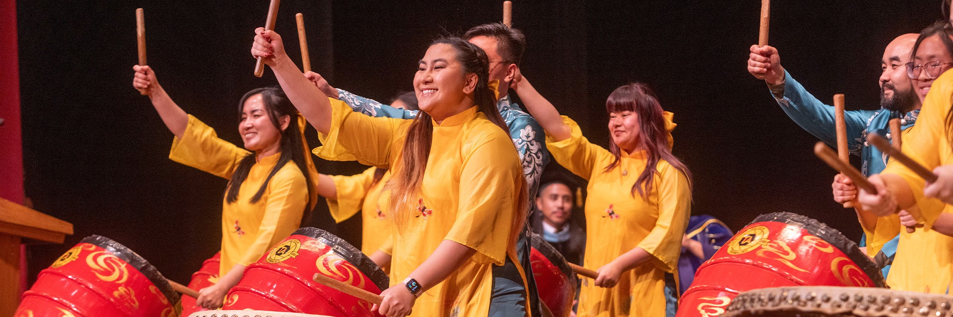 Group of performers on stage playing large red drums, wearing bright yellow traditional-style outfits and smiling while raising drumsticks in unison during a cultural performance.