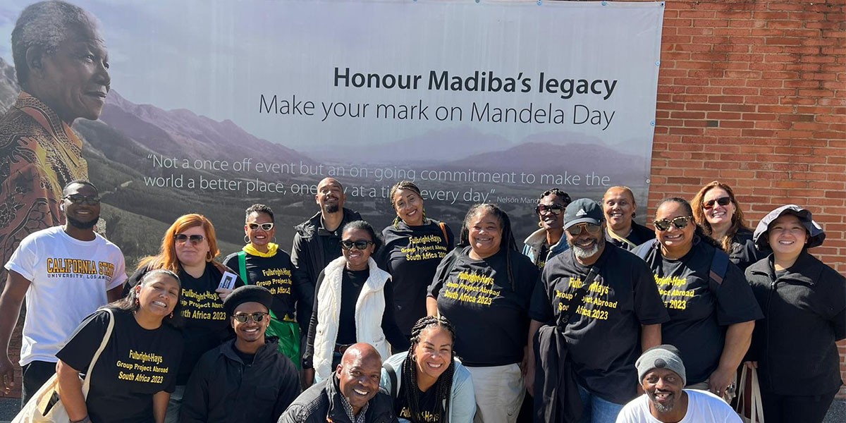 Group of students and faculty in Africa in front of poster that reads Honour Madiba's legacy, make your mark on Mandela Day as part of theFulbright Hays Group Project