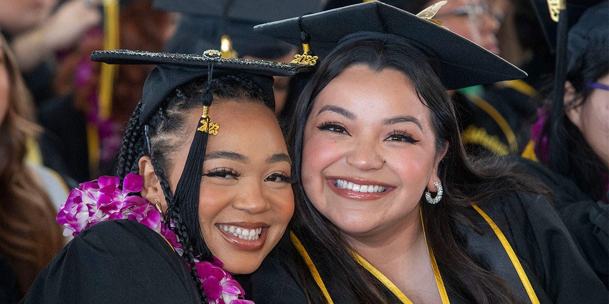 Two graduates cheek to cheek at commencement ceremony wearing graduation robe