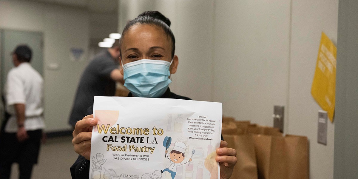 Staff person wearing a facial covering, holding up a "Welcome to the Cal State LA Food Pantry" sign.