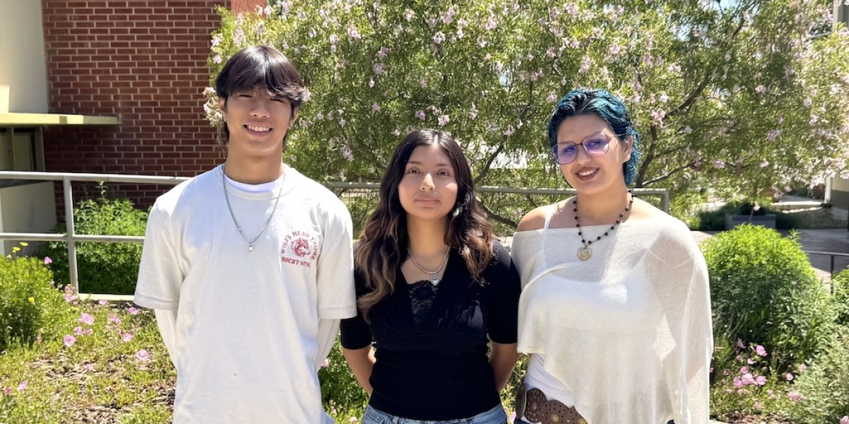 Three young people stand side by side outdoors, smiling at the camera in a sunny garden with blooming flowers and a brick building in the background.
