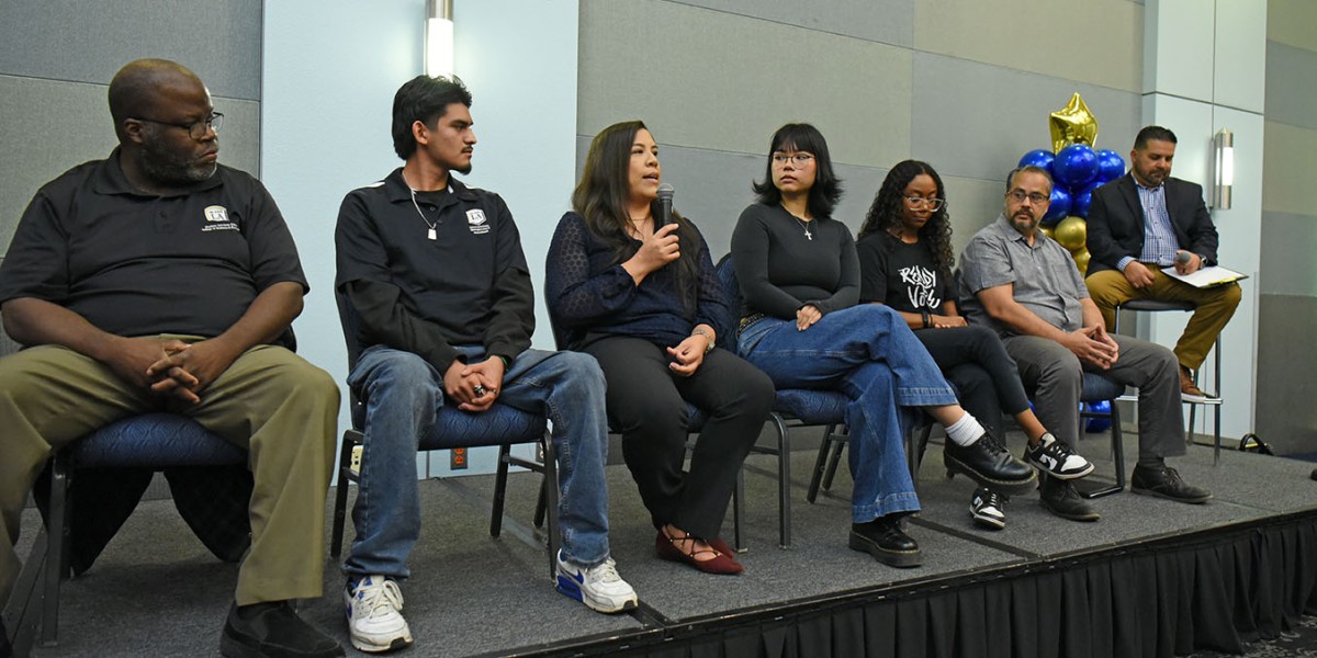 A panel of six people sit on an elevated stage. A moderator sits beside them on a tall stool.