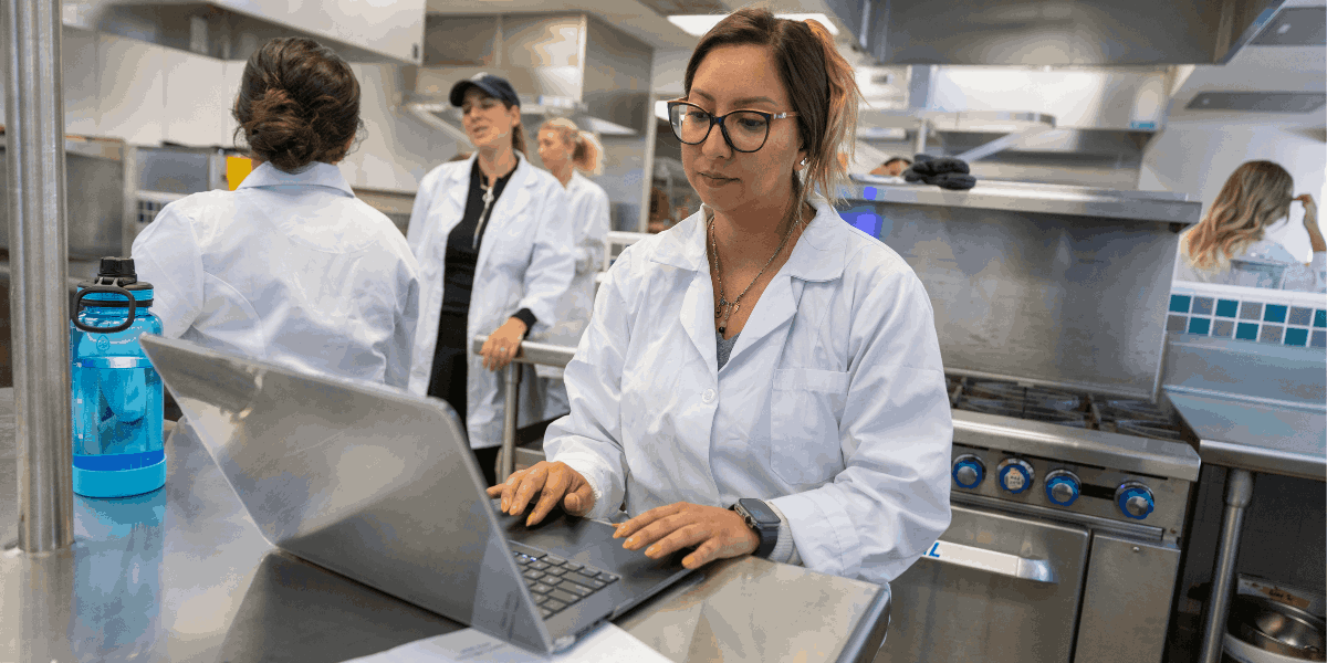 Nutritional science student in industrial kitchen using laptop to input data. Industrial stove. Students.