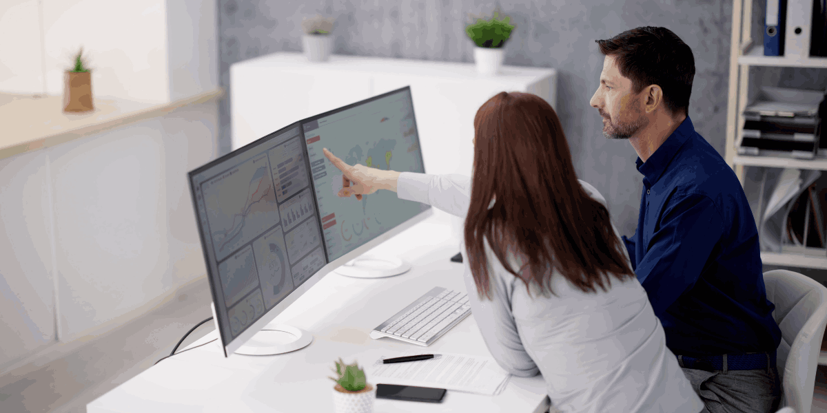 Two individuals are in office looking at desktop computer that displays dashboards.