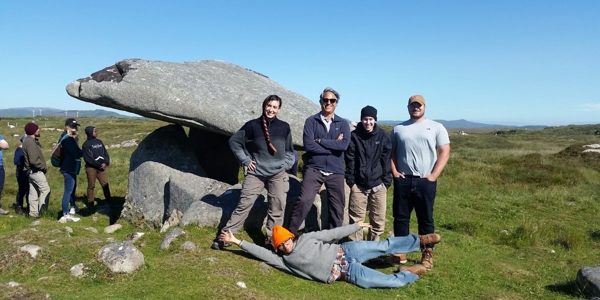 four people posing in front of a rock. Male student posing on the ground. Landscape is green 