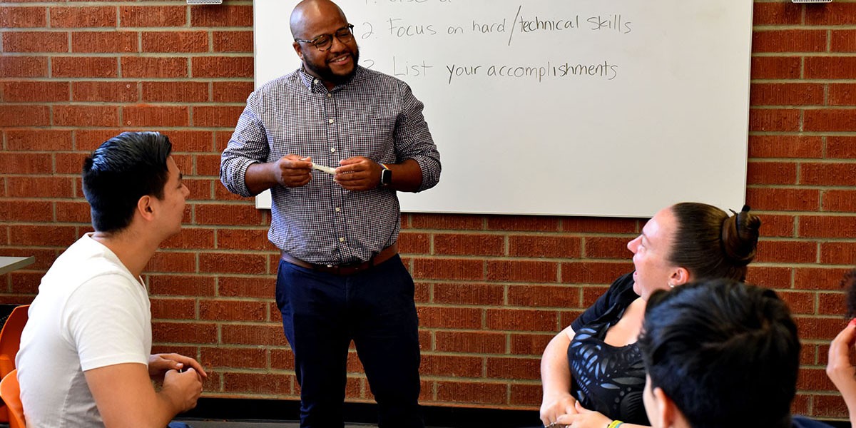 Career advisor stands next to a dry erase board, chatting with students who are seated.