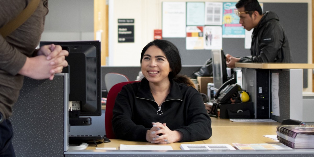 Student employee sits at office desk, smiling at a person standing at the counter.