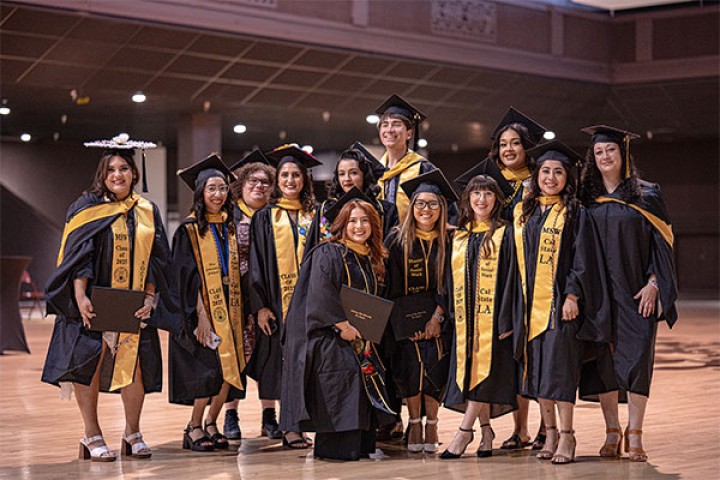 Thirteen Cal State LA MSW graduates from the Class of 2025 stand indoors, smiling in black gowns and yellow stoles.