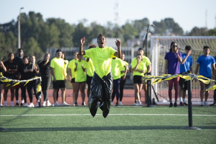 A person with a trash bag tied around their legs hopping down an outdoor grassy field, while spectators watch from behind