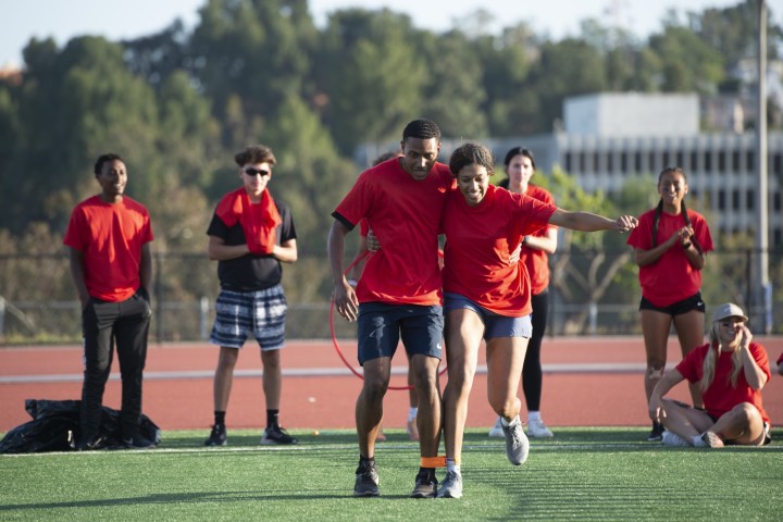 Two students tied together at their legs make their way down a grassy field.