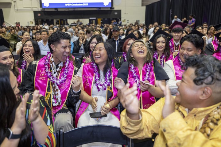 Graduating students wearing pink “Class of 2025” stoles and leis sit in a crowded auditorium, laughing and clapping together during a celebratory ceremony.
