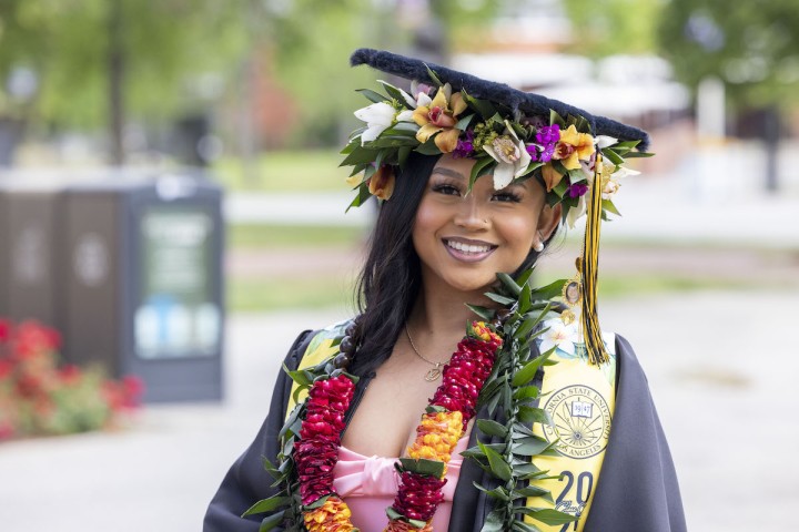 A smiling graduate wearing a decorated cap with flowers, multiple leis, and a Cal State LA stole poses outdoors in cap and gown.