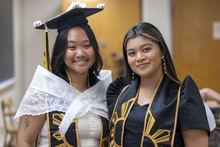 Five people posing together in front of a College of Ethnic Studies backdrop, including a graduate wearing a stole, with yellow and black balloons on the side.