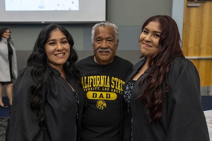 One person standing in between two students who are wearing graduation gowns.