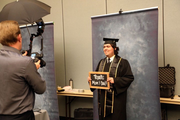 Graduate in cap and gown posing for a professional portrait while holding a sign that reads ‘Thanks Mom & Dad!’ during a Grad Fair photo session.