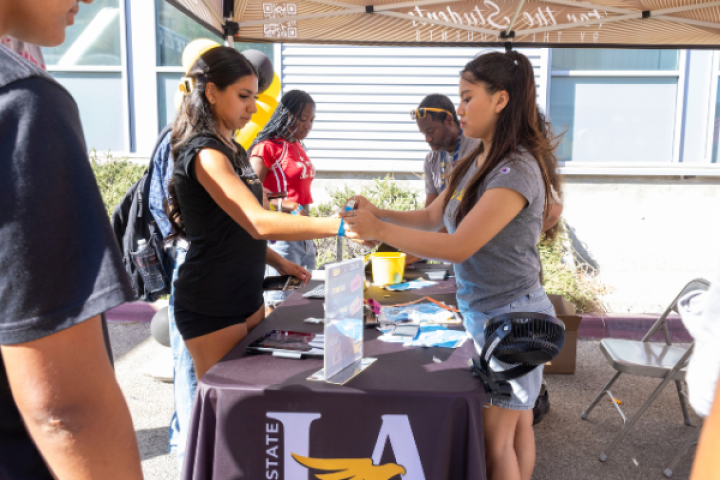 Cal State LA students participate in an outdoor campus activity, with a student staff member assisting an attendee at a table under a canopy.