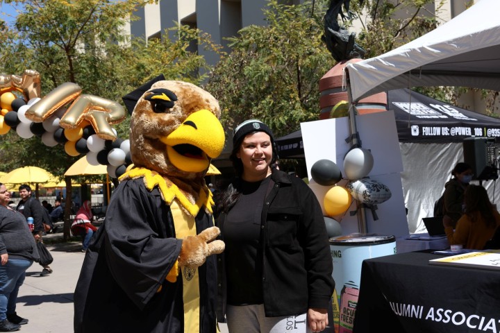 Student posing for a photo with Eddie the Eagle mascot wearing a graduation cap and gown at an outdoor Grad Fair event with tents and balloons on campus