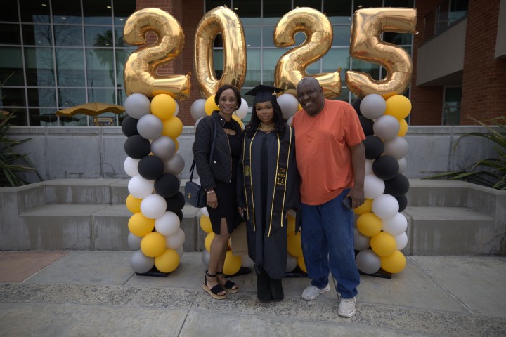 Graduate in cap and gown standing with two guests in front of gold ‘2025’ balloons and black-and-gold balloon columns at Grad Fair