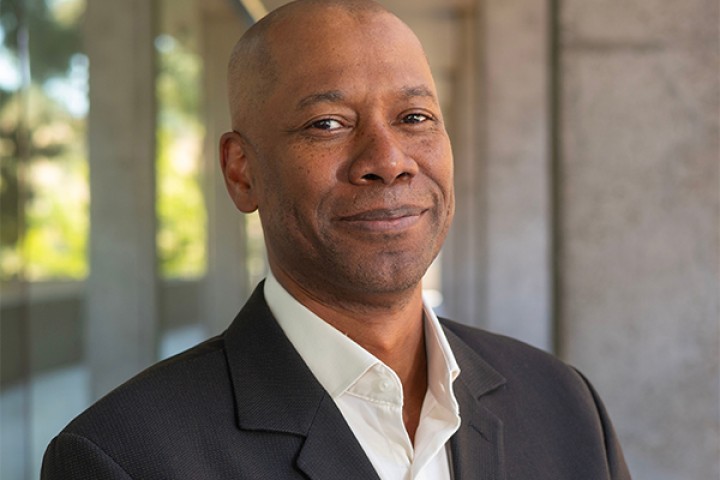 Dean James Ford smiles at the camera while wearing a dark suit jacket and white shirt, photographed outdoors on the Cal State LA campus.