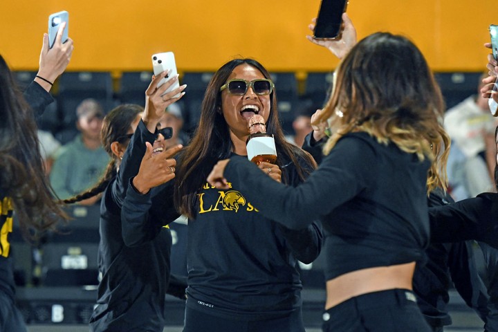 A student wearing sunglasses indoors cheers while holding a mic, surrounded by other students holding their smart phones in the air.