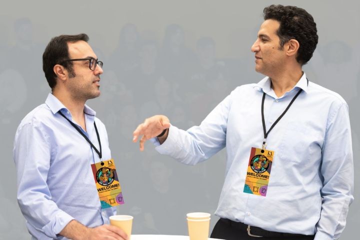 Two men wearing conference badges stand facing each other, talking and gesturing, while holding coffee cups against a neutral background.