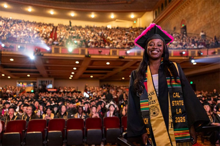 A smiling graduate in a cap and gown stands in front of a large audience during commencement.