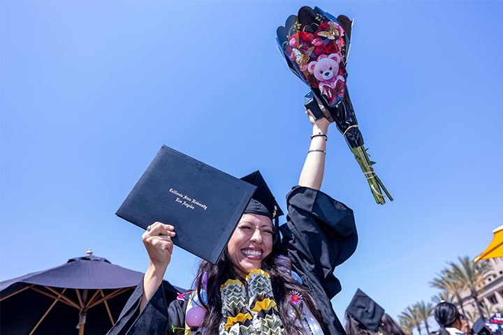Smiling graduate in cap and gown holds up a diploma cover and bouquet while celebrating outdoors after commencement.