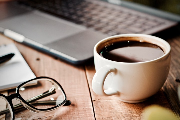 A cup of coffee on a table next to an open laptop computer and a pair of glasses.