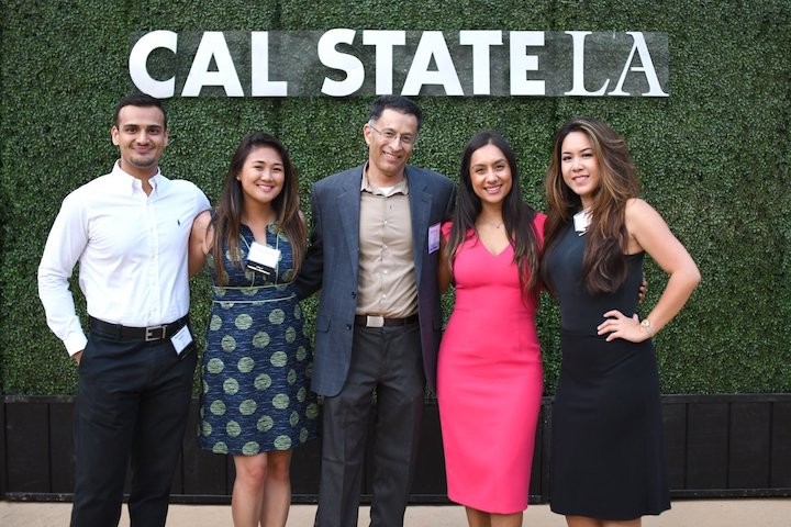 Five smiling alumni posing in front of Cal State LA banner