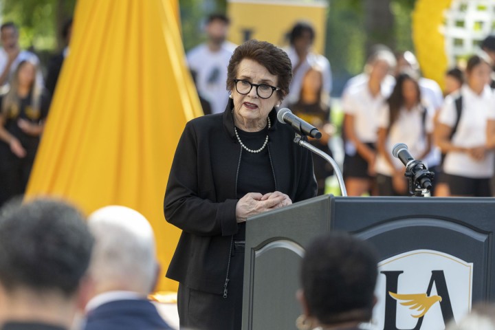 Speaker delivers remarks at a podium during an outdoor campus event, with attendees gathered in the background at Cal State LA. 