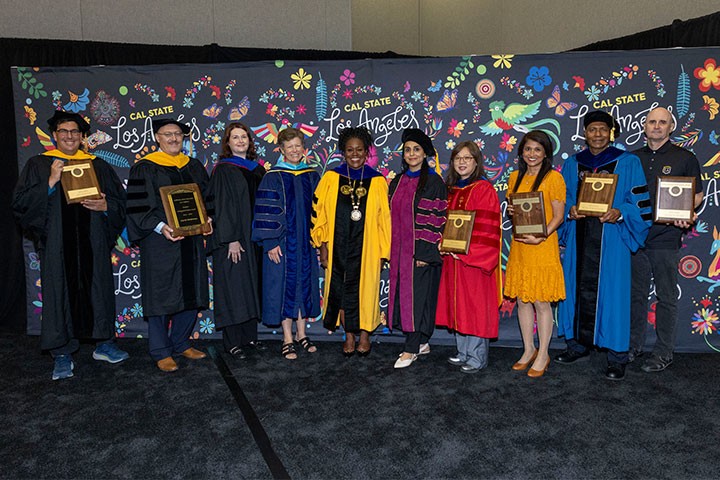 A group of ten Cal State LA faculty and campus leaders stand together on a stage in academic regalia, with several individuals holding award plaques. They pose in front of a black backdrop featuring colorful floral artwork and repeated Cal State Los Angeles logos during a faculty recognition ceremony.