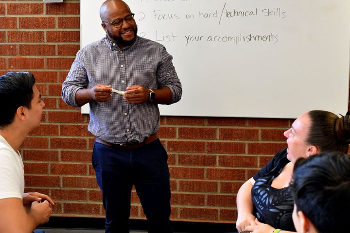 Career advisor stands next to a dry erase board, chatting with students who are seated.