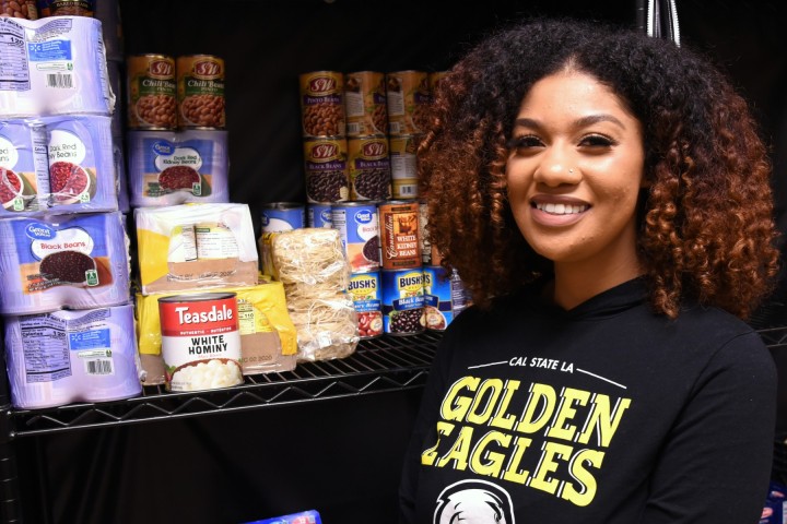 A person smiling, wearing a Golden Eagles sweatshirt, standing in front of shelves stocked with non perishable food.