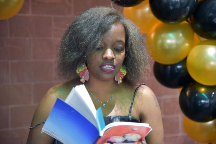 Student reading from a book at an event, balloons in the background.