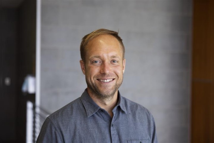 Portrait of a smiling man in a button-down shirt standing indoors against a neutral gray wall background.