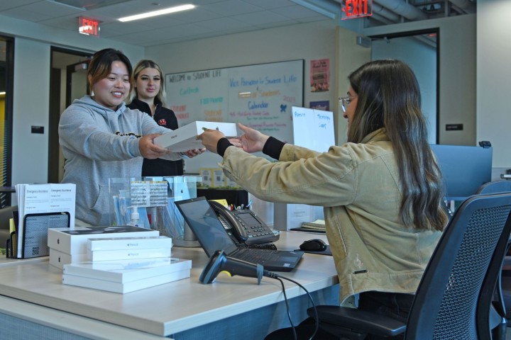 A student smiles while picking up an iPad from a staff member sitting at a desk.