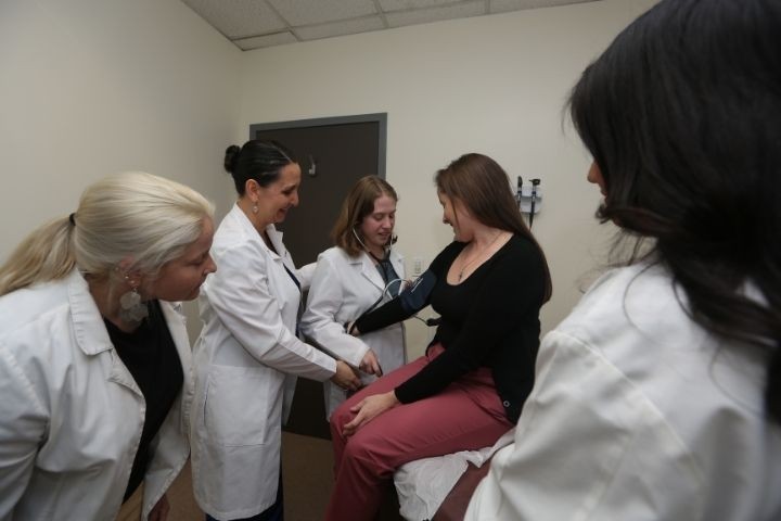 Four females in white coats. One student with stethoscope examining another female 