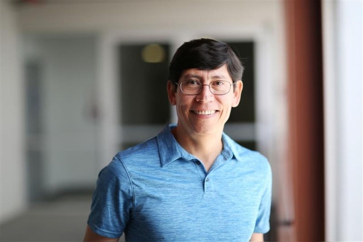 Portrait of a smiling man in a blue polo shirt and glasses standing in a softly lit indoor corridor with a blurred background. 