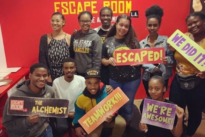 Group of students and staff in an escape room, smiling and holding up signs.
