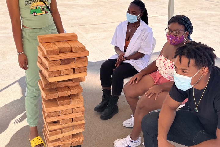 Three students wearing facial covering and sitting and one students standing, surrounding a large Jenga game.