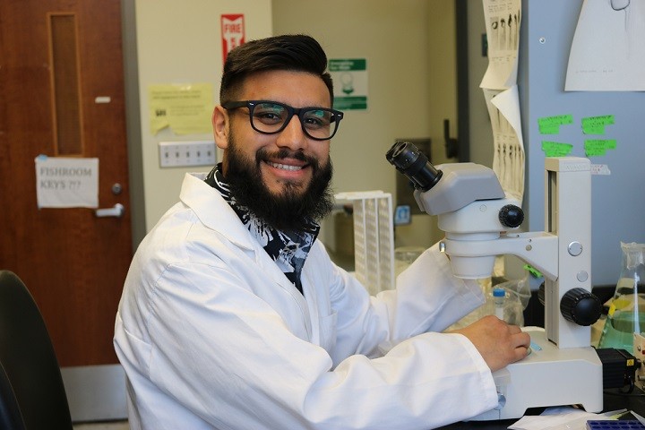male student with a beard, glasses and white lab coat smiling at the camera. He is holding a microscope in a lab