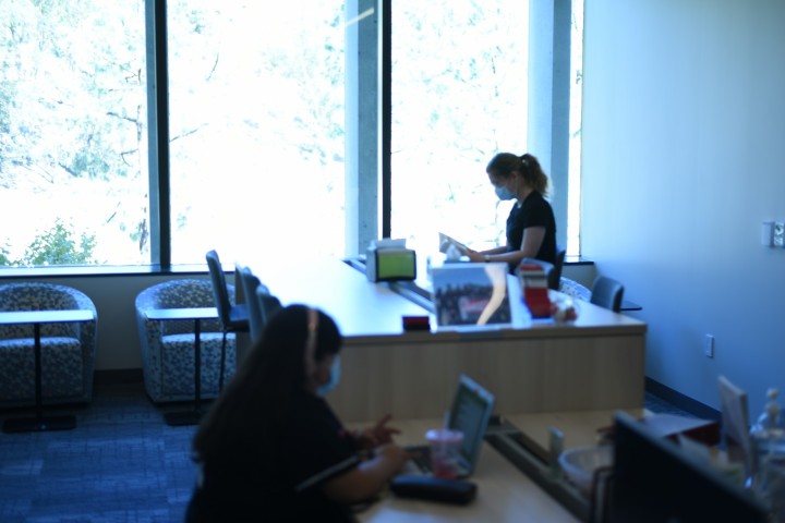 Student with headphones sits at table reading. Student in the background sits at tall table reading. 