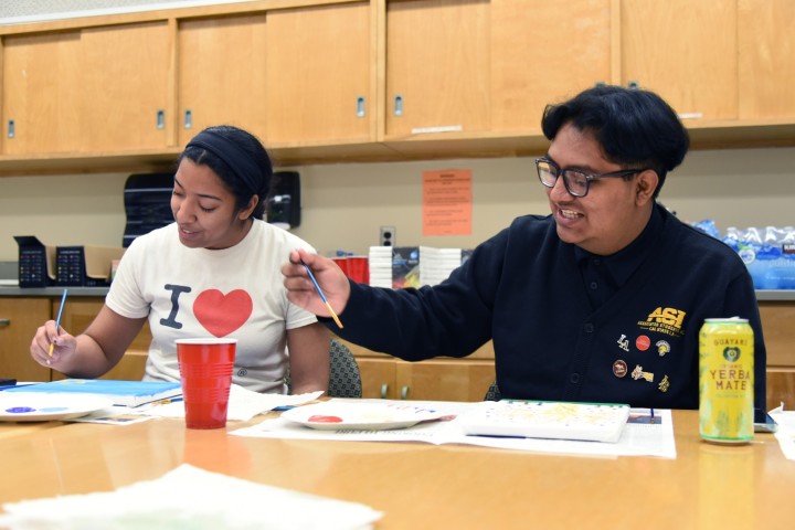 Two students seated at a table smiling while painting.