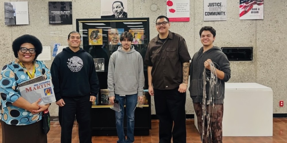Dr. Tarisi Vunidilo and students stand together in front of a MLK display, with posters about justice, equity, and community visible behind them
