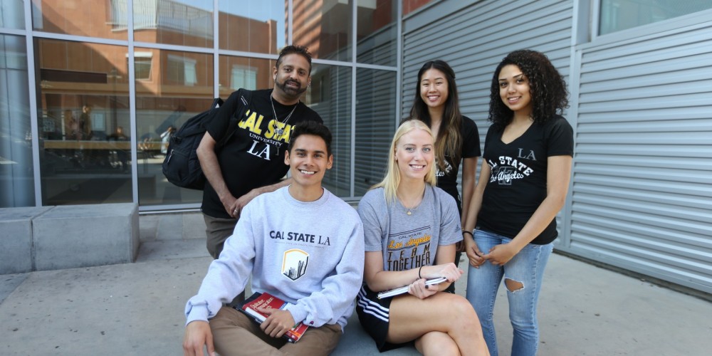 five students standing in front of a building with glass. Two students sitting down and three right behind