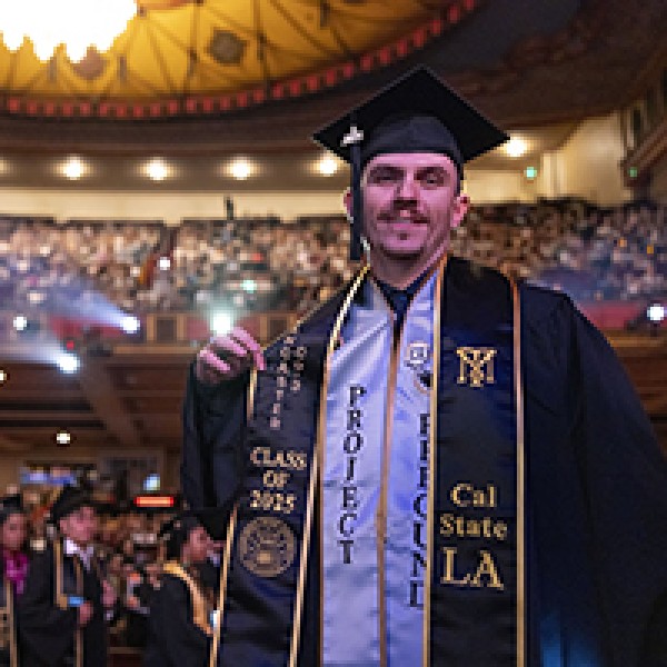 A student wearing graduation attire smiles, an auditorium full of people behind him.