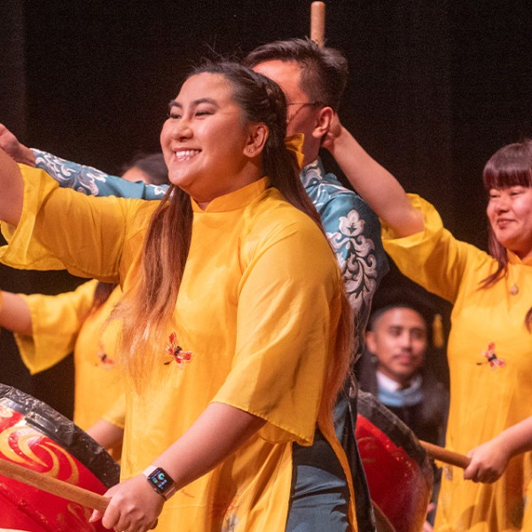 Group of performers on stage playing large red drums, wearing bright yellow traditional-style outfits and smiling while raising drumsticks in unison during a cultural performance.