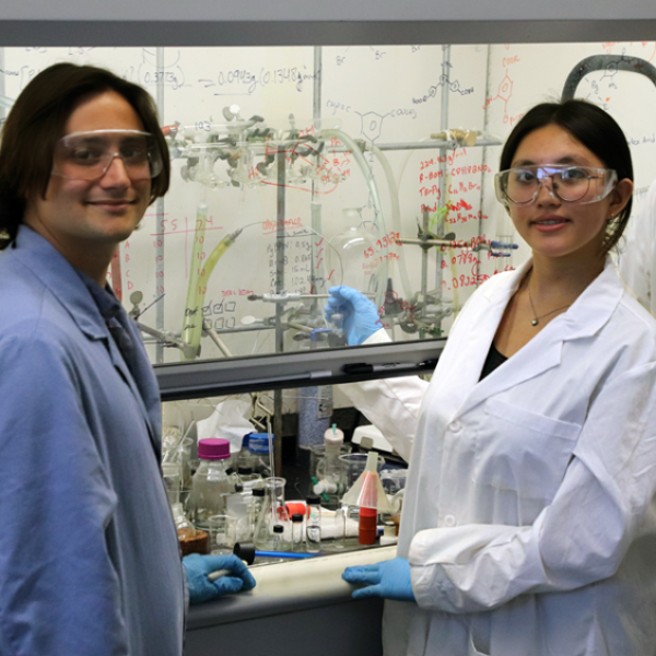 students in lab with coat gloves and goggles experimenting behind glass with equations written 