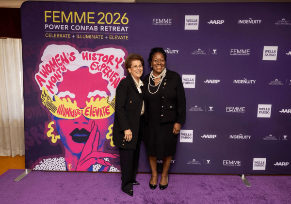 Two leaders pose together at the FEMME 2026 Power Confab Retreat in front of a purple backdrop with “Women’s History Experience” artwork.